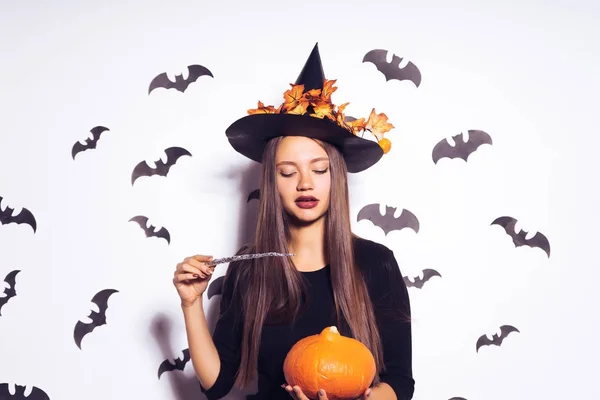 Una joven misteriosa vestida con una bruja para Halloween, sosteniendo una calabaza naranja y una varita mágica, evoca —  Fotos de Stock