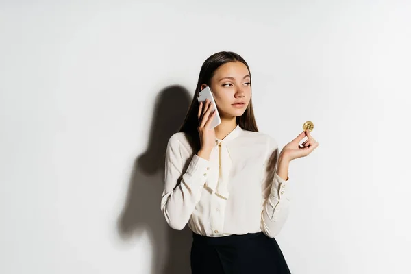 Уверенная успешная девушка в белой рубашке держит в руке золотой биткойн и говорит по телефону о криптовалюте — стоковое фото