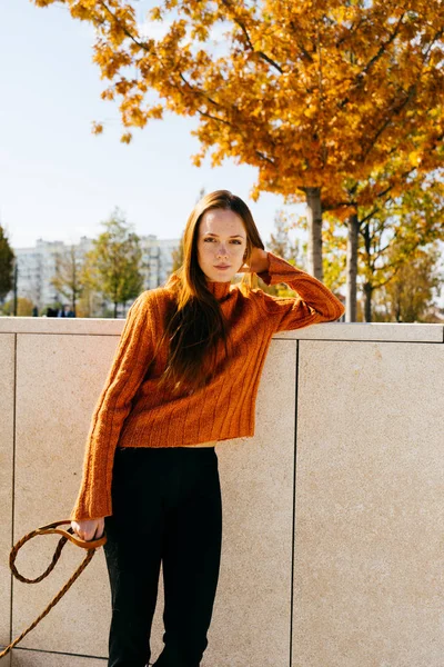 Linda menina ruiva doce em um suéter laranja na moda posando por uma cerca leve em um parque de outono — Fotografia de Stock