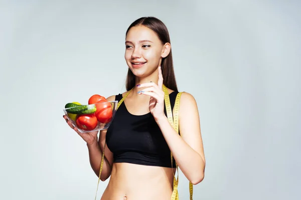 Jovem menina feliz em tops esportivos segue sua figura, segura um prato com pepinos e tomates em sua mão — Fotografia de Stock