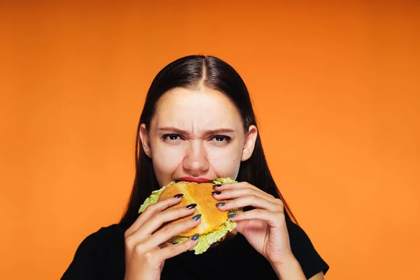 ハンバーガーを食べて、オレンジ色の背景に怒っている女の子 — ストック写真