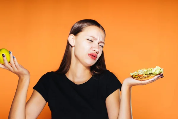 Naštvaná holka na oranžové pozadí kvákání burger a apple ve svých rukou — Stock fotografie