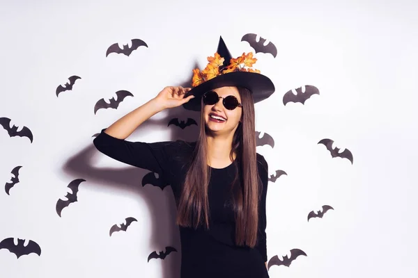 Beautiful laughing gothic girl in the image of a witch, celebrating halloween, wearing black sunglasses, a big black hat decorated with yellow leaves — Stock Photo, Image