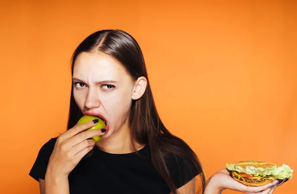 Злая молодая девушка сидит на диете, ест полезное зеленое яблоко и держит в руке вредный гамбургер — стоковое фото