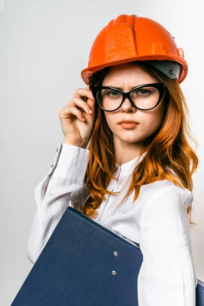 Uma jovem garota séria com óculos e um capacete protetor laranja mantém uma pasta com documentos importantes — Fotografia de Stock