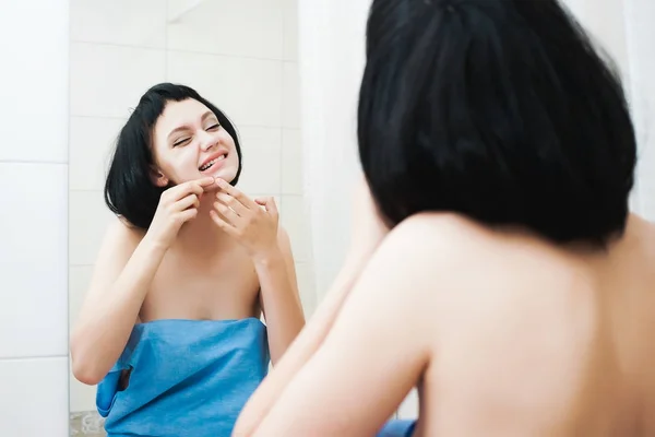 Молода красива дівчина з чорним коротким волоссям в синьому рушнику дивиться у дзеркало і стискає прищик на обличчі — стокове фото
