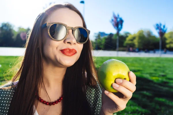 Krásná mladá dívka v sluneční brýle s červenými rty jíst jablko v parku — Stock fotografie