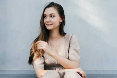 bej renkli elbiseli güzel gülümseyen kız beyaz bir duvar tarafından oturur ve uzak görünüyor