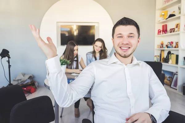 Młodego człowieka sukcesu, w białej koszuli siedzi w biurze, śmieje się, za nim pracują 2 piękne dziewczyny — Zdjęcie stockowe