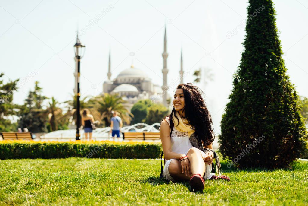 A bright Turkish girl sits on the grass on the background of the Blue Mosque in Istanbul