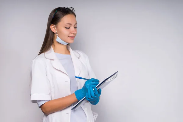 Bella giovane medico ragazza in vestaglia medica bianca scrive qualcosa in una cartella con i documenti — Foto Stock