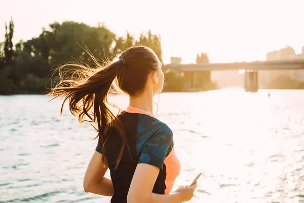 Młoda dziewczyna sportowa z długimi włosami jogging nad rzeką o zachodzie słońca, słuchanie muzyki na słuchawkach — Zdjęcie stockowe