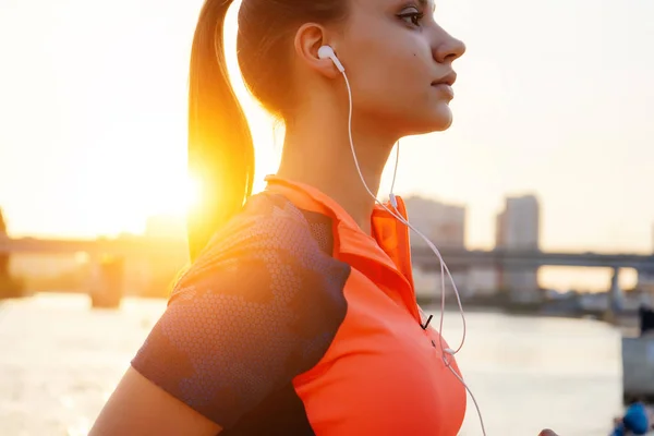 Vackra sport flicka bedriver konditionen på floden, vid solnedgången, lyssna på musik i hörlurar — Stockfoto