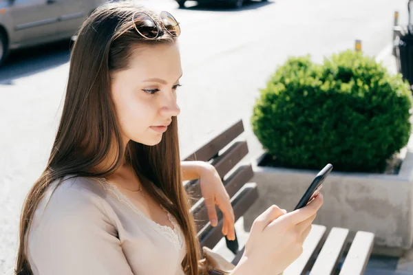 Young beautiful girl in a beige dress and sunglasses sits on a bench outside and looks in her smartphone — Stock Photo, Image