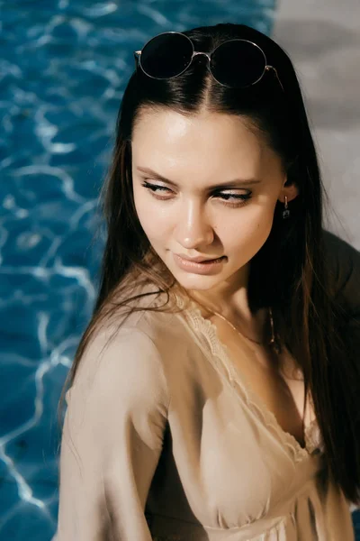 Hermosa mujer con el pelo largo y vestido beige se sienta junto al agua azul en el sol — Foto de Stock