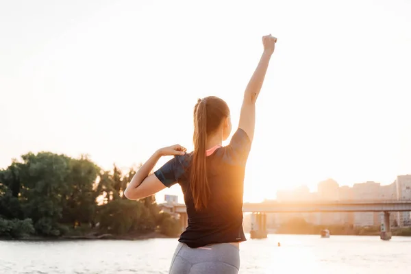 Slim atletisk tjej gör konditionen på floden i solnedgången, lyssna på musik i hörlurar — Stockfoto