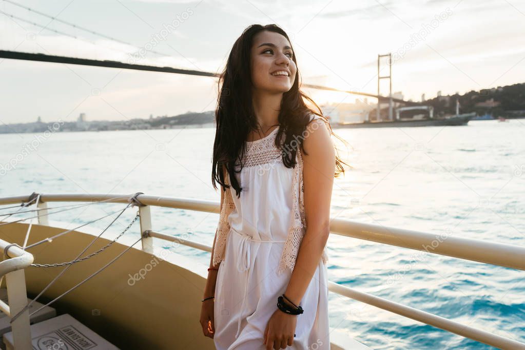 Young girl traveler relaxes while cruising on a yacht, looking at sunset, modern famous bridge on background