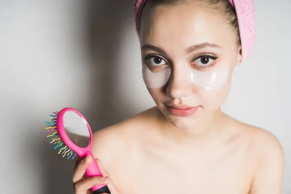 Молодая красивая девушка с розовым полотенцем на голове и силиконовыми пятнами под глазами держит расческу в руке с зеркалом и смотрит в камеру — стоковое фото
