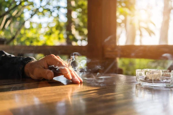 За деревянным столом сидит мужчина, его рука лежит на столе и держит сигарету — стоковое фото