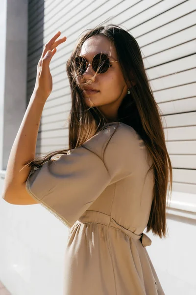 Ragazza dai capelli lunghi con un vestito beige e occhiali da sole che camminano lungo la strada al sole — Foto Stock
