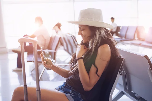 Airport woman in hat  on smart phone at gate waiting in terminal. Air travel concept with young hipster sitting with hand luggage suitcase. Beautiful young mixed race female — Stock Photo, Image