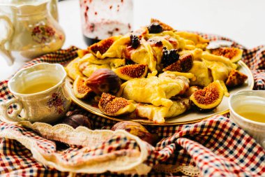 Beyaz bir tablo ortada parlak damalı masa örtüsü, börek ve tatlı incir, çay bardağı ile bir tabak