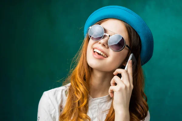 Ragazza dai capelli rossi alla moda con un berretto blu e occhiali da sole che parlano al telefono, sorridono, sembrano felici — Foto Stock