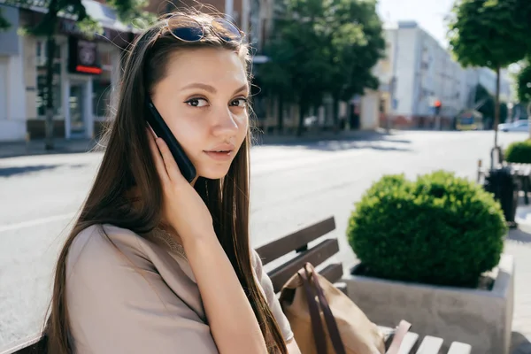 bir şehir sokakta bir bankta oturmuş, telefonda konuşurken ve kameraya bakarak sevimli kız