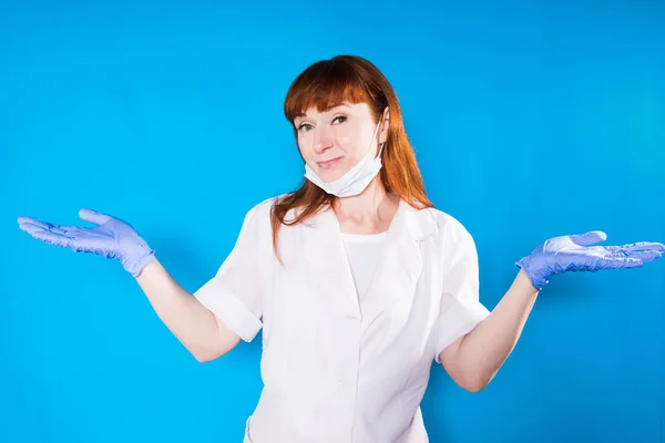 Rödhåriga sjuksköterska i en mask sprida hennes händer till sida och ser leende på kameran, isolerad på en blå bakgrund — Stockfoto