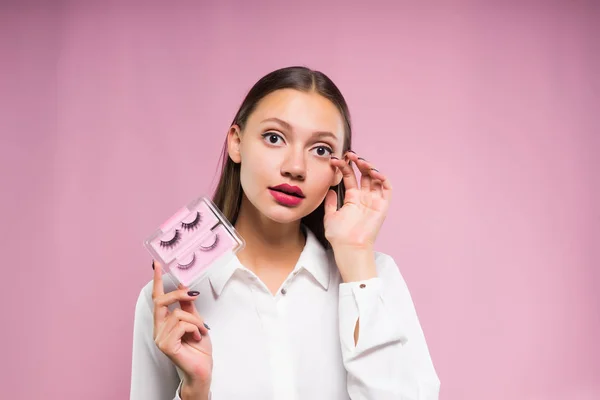 Fille avec de gros yeux tient de faux cils et regarde dans la caméra, isolé sur fond rose — Photo