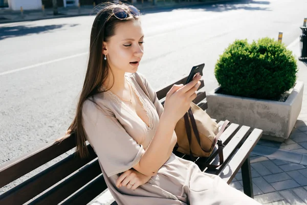 Sexy chica joven en un vestido beige y con gafas de sol se sienta en un banco fuera, mira en su teléfono inteligente — Foto de Stock