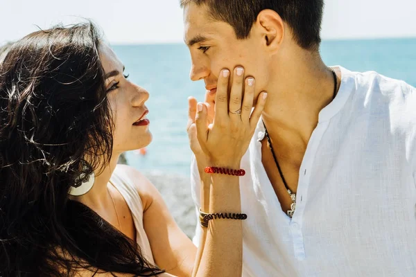 Ung vacker flicka håller ansiktet av sin pojkvän och ser försiktigt in i ögonen på bakgrunden av havet — Stockfoto