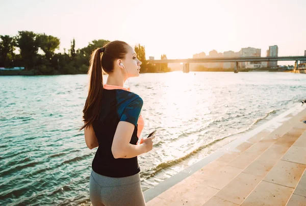 Flicka i sport kostym jogging på floden vallen lyssnar på musik i hörlurar — Stockfoto