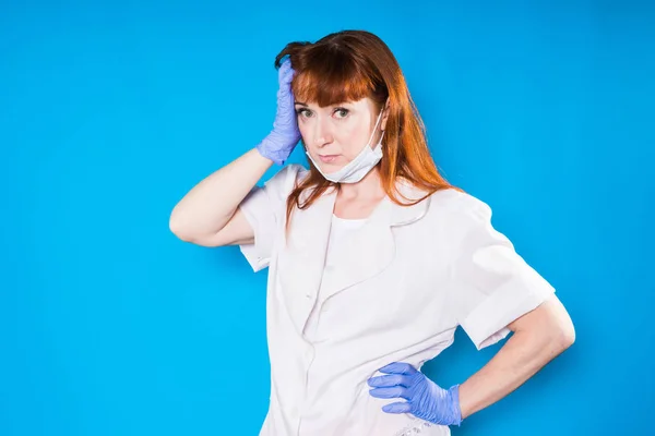 Müde nach der Arbeit hält eine Krankenschwester in Handschuhen ihren Kopf und blickt in die Kamera, isoliert auf blauem Hintergrund — Stockfoto
