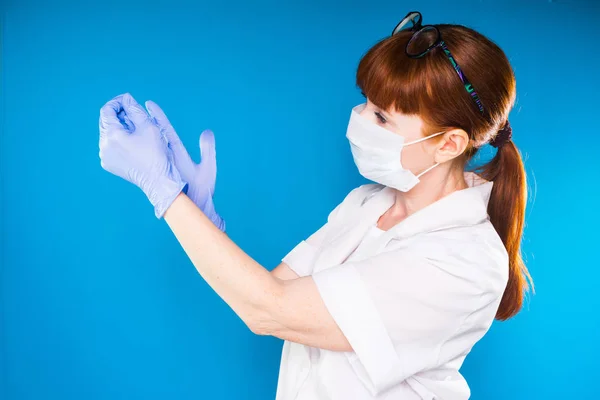 Kvinna sjuksköterska i medicinska kläder och bära en mask sätter på gummihandskar, isolerad på en blå bakgrund — Stockfoto