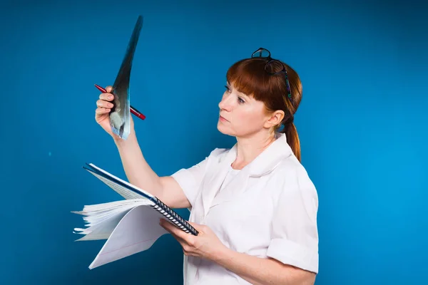 Rothaarige Frau in weißem Gewand betrachtet ein Röntgenbild und hält ein Papier in der Hand, isoliert auf blauem Hintergrund — Stockfoto