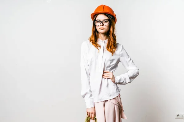 Dai capelli rossi ragazza fiduciosa in occhiali e un casco protettivo arancione, in una camicia bianca posa contro una parete bianca — Foto Stock