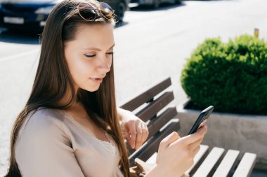 bej renkli elbiseli genç çekici kız sokakta bir bankta oturur, onun smartphone görünüyor