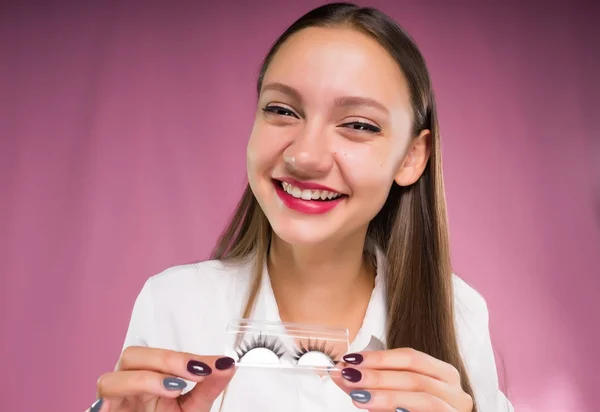 Beautiful laughing girl holding a package with false eyelashes, behind a pink background — Stock Photo, Image