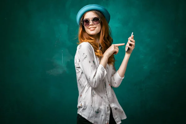 Affascinante ragazza elegante in un berretto blu e occhiali da sole sta tenendo uno smartphone e punta ad esso con il dito, sorridendo — Foto Stock