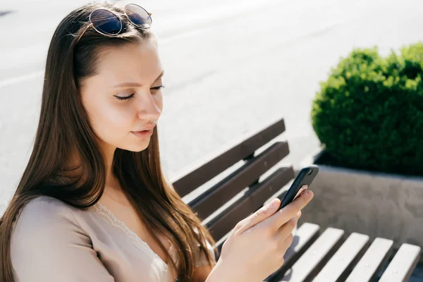 Bella ragazza attraente in un abito beige si siede su una panchina di legno, guarda nel suo smartphone — Foto Stock