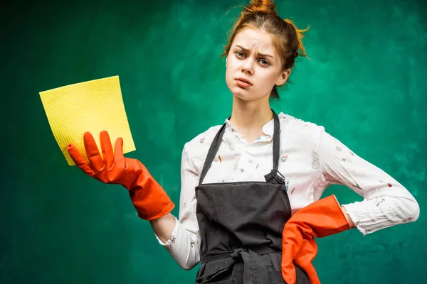 Sad young girl in a black apron, wearing orange rubber gloves holding a yellow rag — Stock Photo, Image