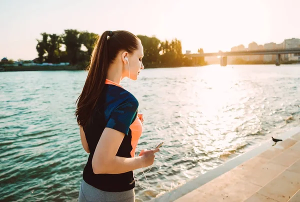 Unga fitness tjej jogging nära floden vid solnedgången, lyssna på musik i hörlurar — Stockfoto