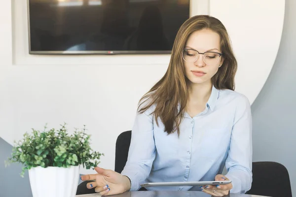 Успешная уверенная девушка в очках и синей рубашке работает в офисе, глядя на планшет — стоковое фото