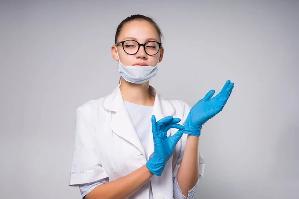 Ung kvinna läkare bär glasögon och en vit kappa, sätta på blå medicinska handskar, listigt tittar åt sidan — Stockfoto