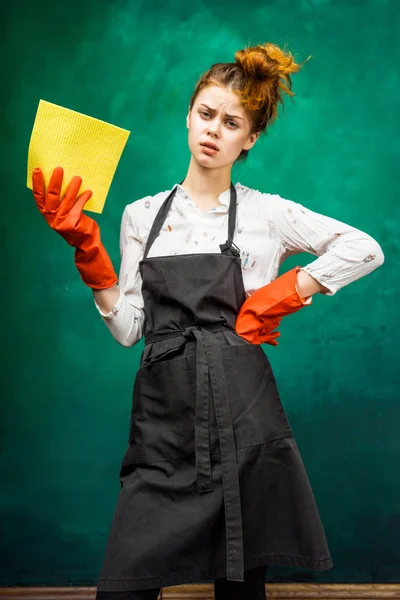 A tired young girl in a black apron, wearing orange rubber gloves holds a yellow rag in her hand — Stock Photo, Image