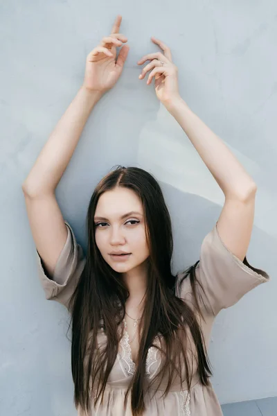 Menina sexy atraente em um vestido bege posando contra uma parede leve, seus braços levantados — Fotografia de Stock