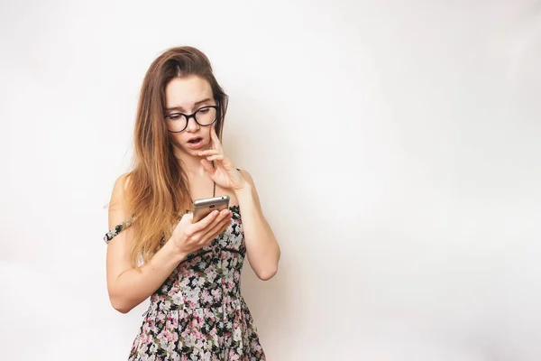 Verbaasd gezicht vrouw geïsoleerd, in glazen en een trendy jurk, kijkt naar haar smartphone — Stockfoto