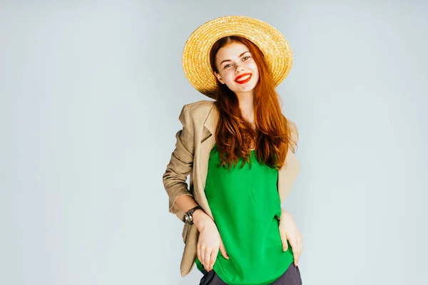 happy red-haired girl in a straw hat, with red lipstick and in a green jacket smiles and poses