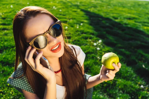 嫌そうな顔の女性のスマート フォンを使用して、青リンゴを手で保持して、緑の草の上に座っています。 — ストック写真
